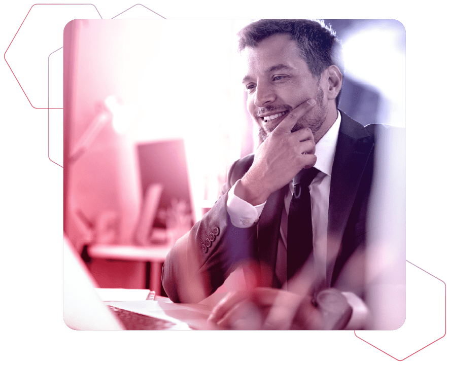 Ein lächelnder Mann im Anzug sitzt im Büro vor seinem Laptop und hält sich eine Hand an sein Kinn auf rot-blauem Hintergrund