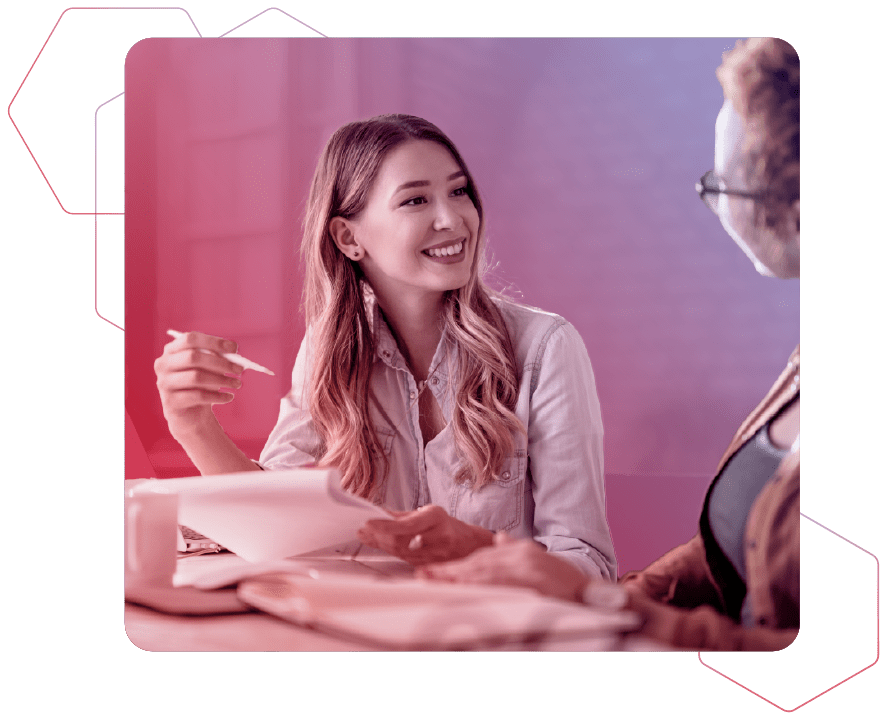 Zwei Frauen die sich im Büro miteinander an einem Tisch unterhalten und Unterlagen vor sich liegen haben auf rot-blauem Hintergrund