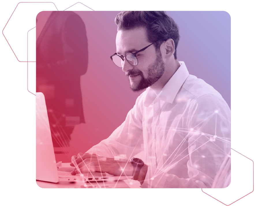 Ein junger bärtiger Mann mit Brille und einem weißen Hemd arbeitet an seinem Laptop auf rot-blauem Hintergrund