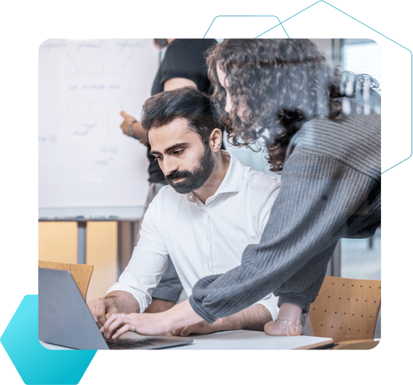 Im Vordergrund erklärt eine Frau einem Mann etwas an einem Laptop, während im Hintergrund ein Mann ein Whiteboard beschriftet